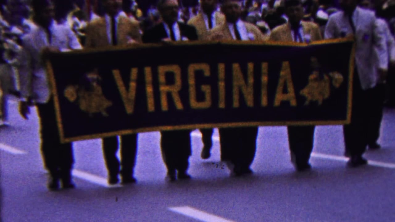 In metadata-supported 1963 Indiana parade footage, men in light-colored jackets carry a large black and gold VIRGINIA banner as spectators fill the background.