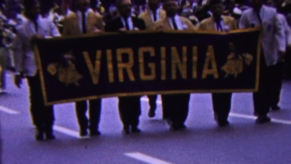 In metadata-supported 1963 Indiana parade footage, men in light-colored jackets carry a large black and gold VIRGINIA banner as spectators fill the background.