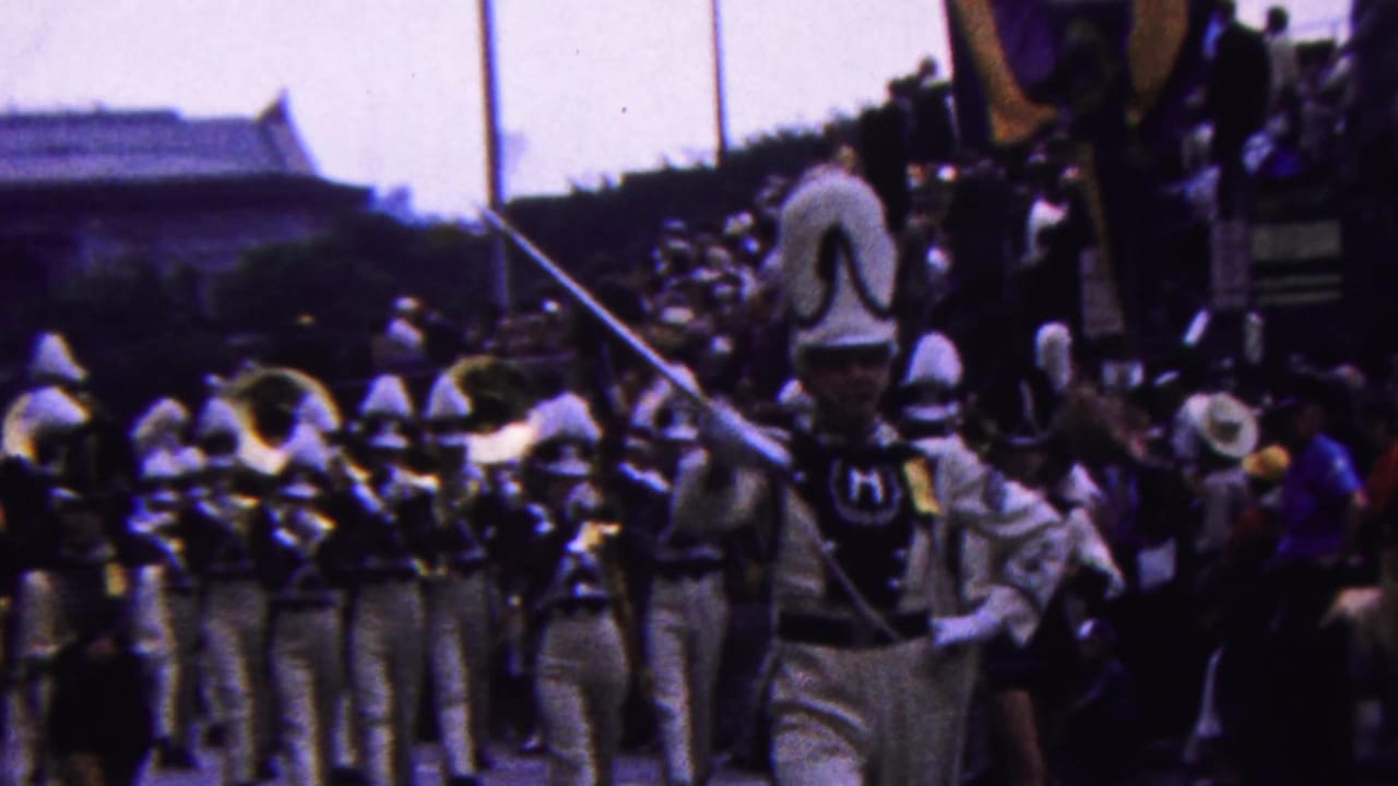 A white-uniformed marching band with tall hats and brass instruments advances along a crowded parade route in metadata-supported 1963 Indiana footage.