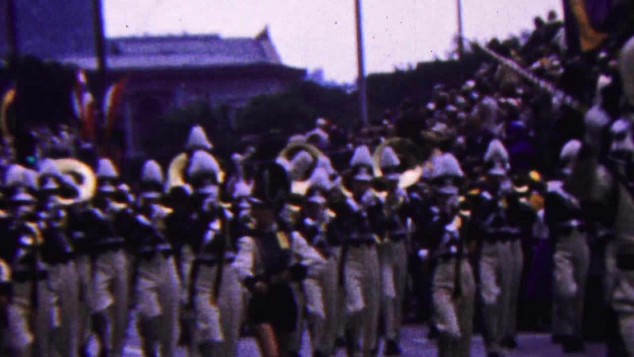 Band members in white uniforms and tall hats continue marching through the parade crowd, with brass instruments and a partially visible banner in the background.