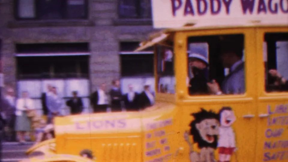 The yellow paddy wagon float fills the frame as passengers ride inside and painted lettering, including Lions Club references, appears along the side during the Indiana parade scene.