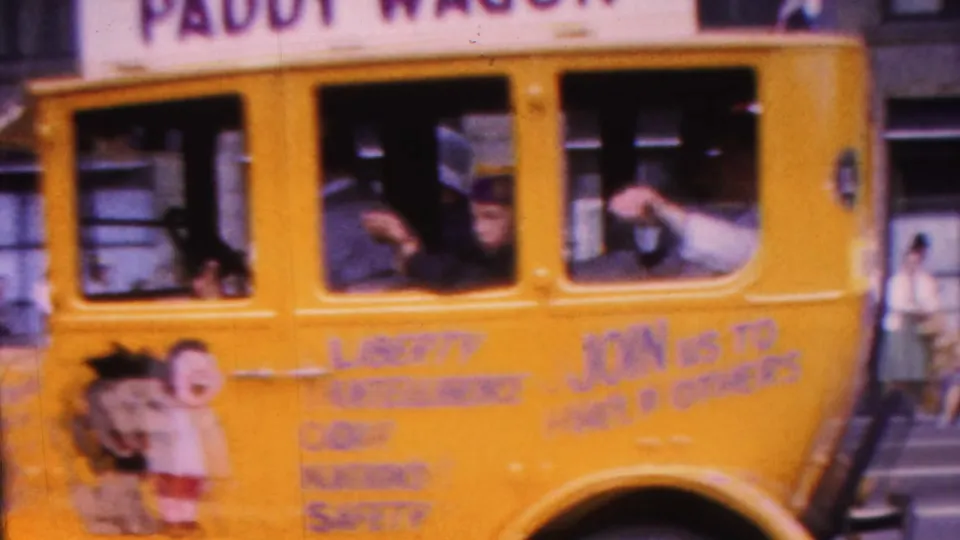 The rear portion of the Lions Club paddy wagon float passes close to camera, showing hand-painted civic slogans, a cartoon lion, and riders waving from the windows.