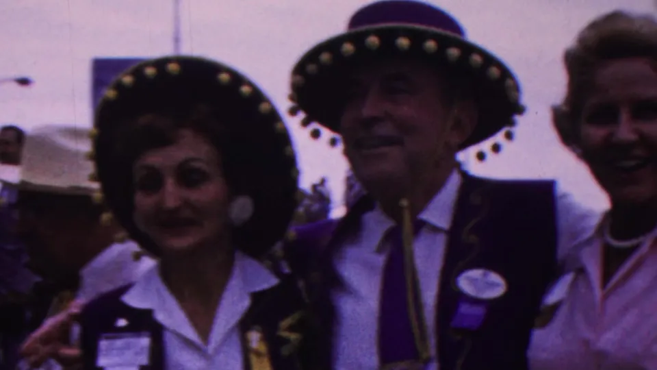Two adult parade participants wearing decorated sombrero-style hats smile toward the home-movie camera, with another woman standing beside them in the supported 1963 Indiana footage.