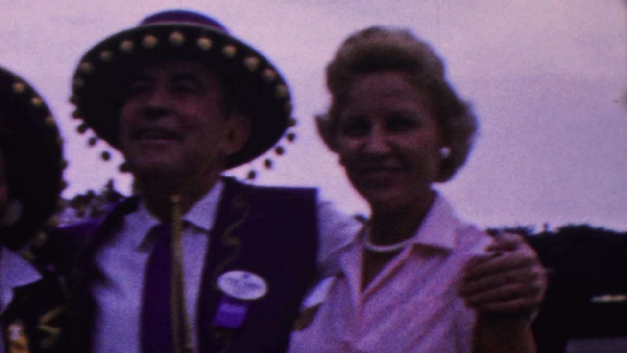 In 1963 Indiana home-movie footage, an adult man in a purple vest and decorated hat stands close to a smiling woman wearing a light blouse and pearl necklace at an outdoor public event.