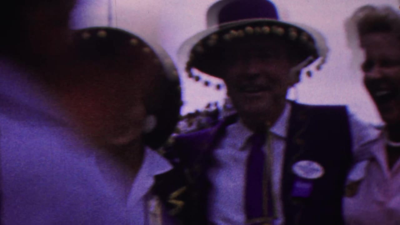 A blurred passerby partly blocks adults in hats and vests posing outdoors.