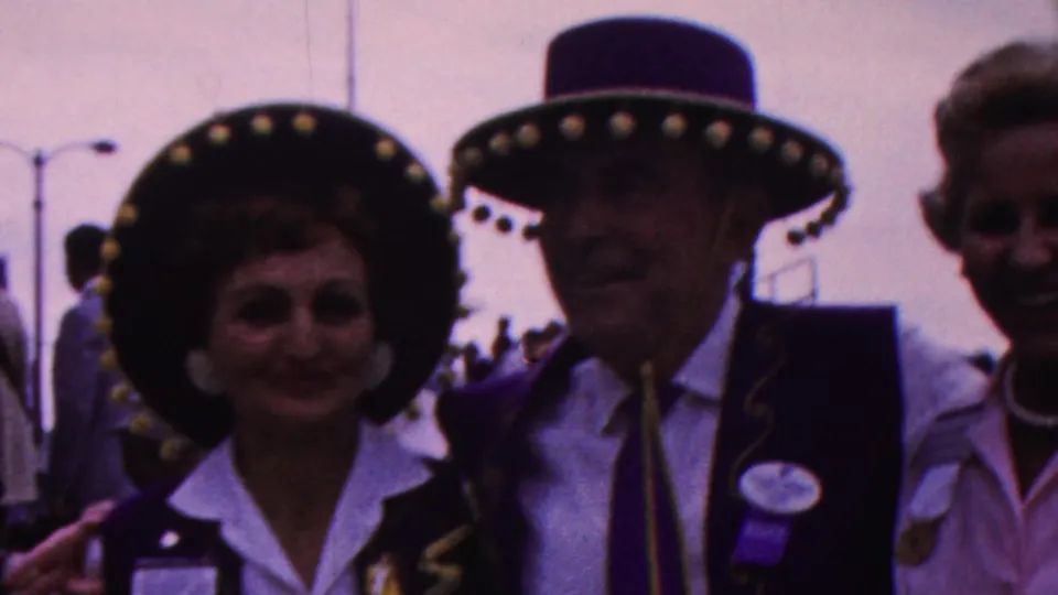 A woman in a decorated wide-brimmed hat and white collared shirt stands beside a man in a matching hat and purple vest, with another smiling woman partly visible behind them.