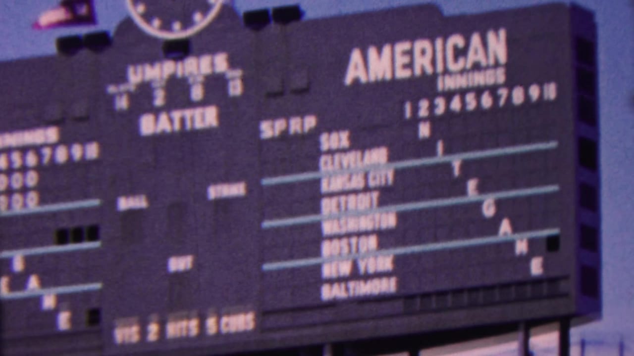 1963 Indiana 8mm frame of a large stadium baseball scoreboard labeled American, with team names, inning numbers, and an analog clock visible above.