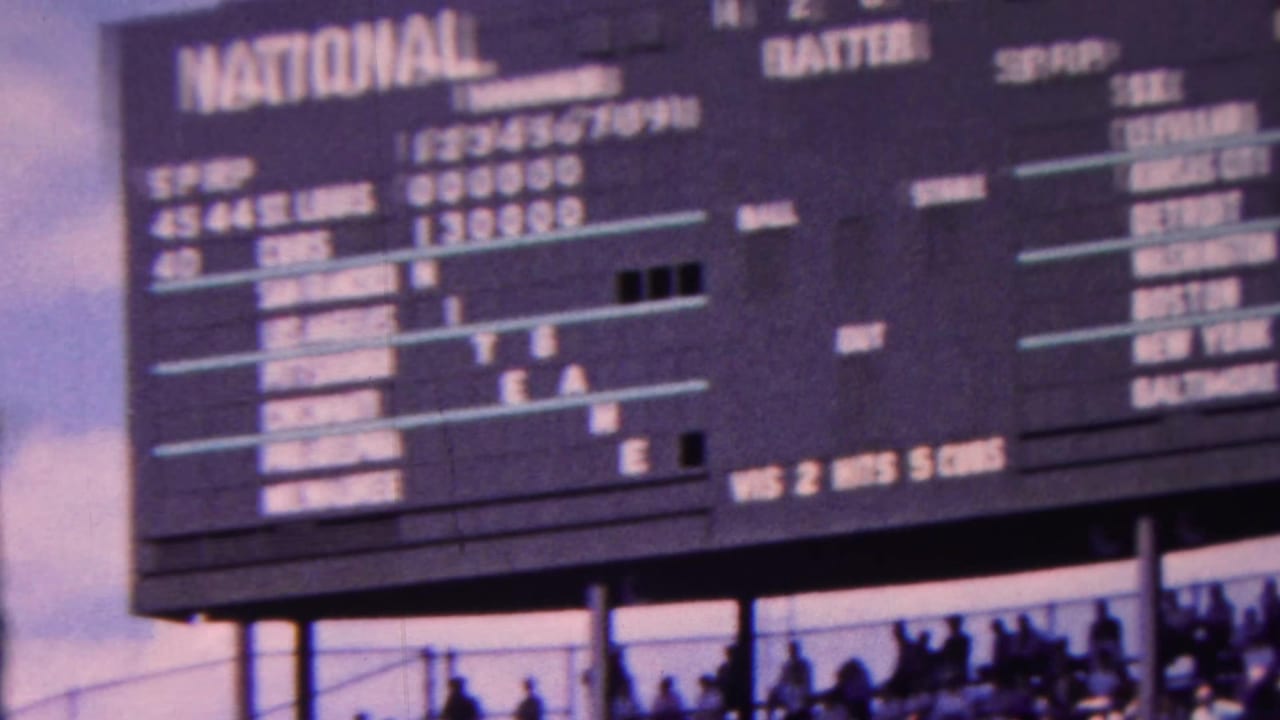 1963 Indiana home-movie frame showing large National and American baseball scoreboard panels with game lines, team names, and silhouetted spectators below.