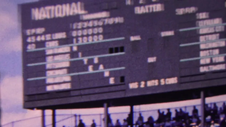 1963 Indiana home-movie frame showing large National and American baseball scoreboard panels with game lines, team names, and silhouetted spectators below.