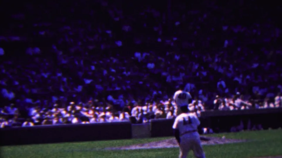 Distant 1963 Indiana 8mm view of a baseball player in a white uniform on the field, with packed stadium seating and a dark outfield wall behind him.