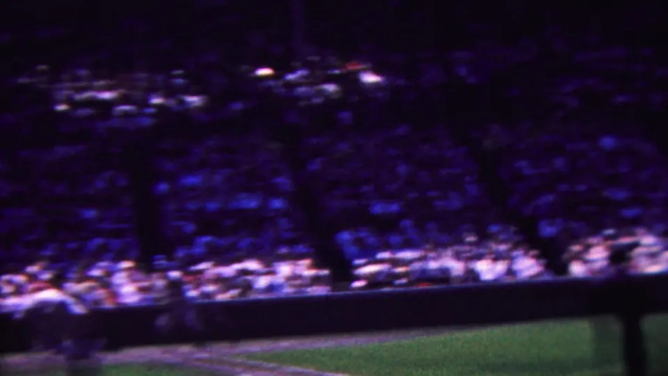 1963 Indiana home-movie frame with motion-blurred stadium spectators, green field, a dark foreground obstruction, and indistinct figures near the playing area.