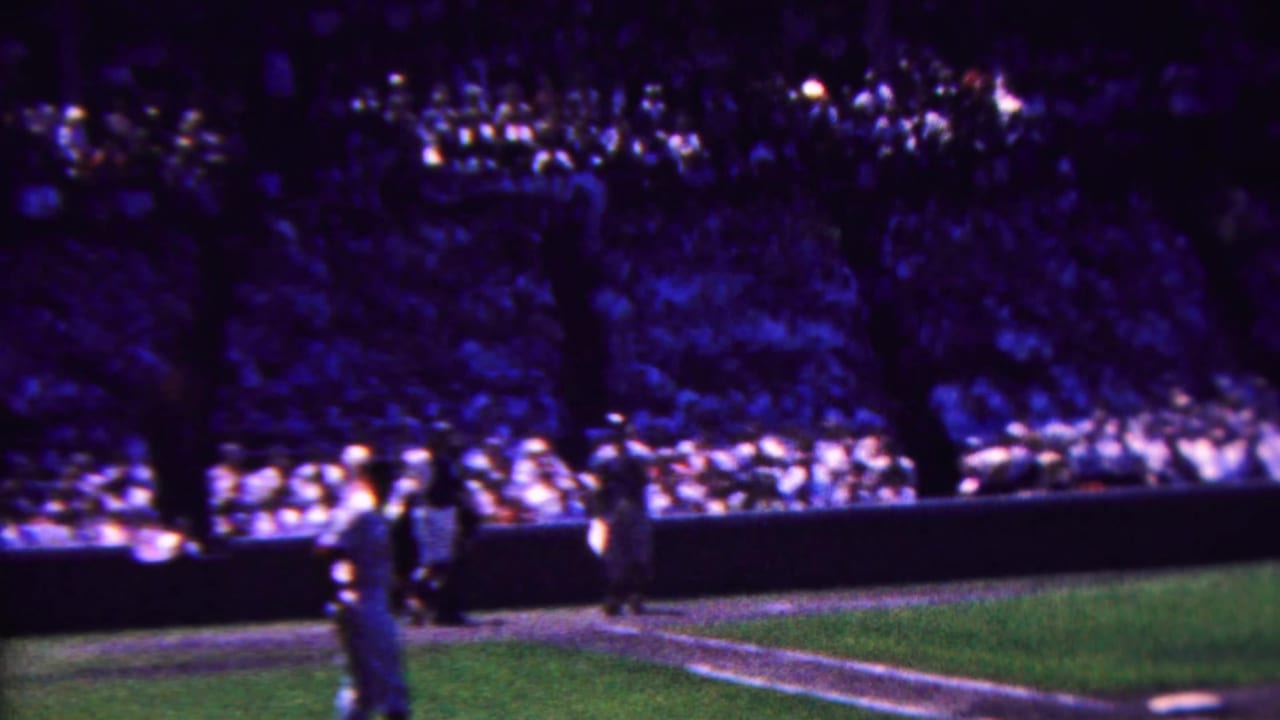 1963 Indiana 8mm home-movie frame showing a crowded baseball stadium, a uniformed player near the field edge, grass, low barriers, and dense spectator seating.