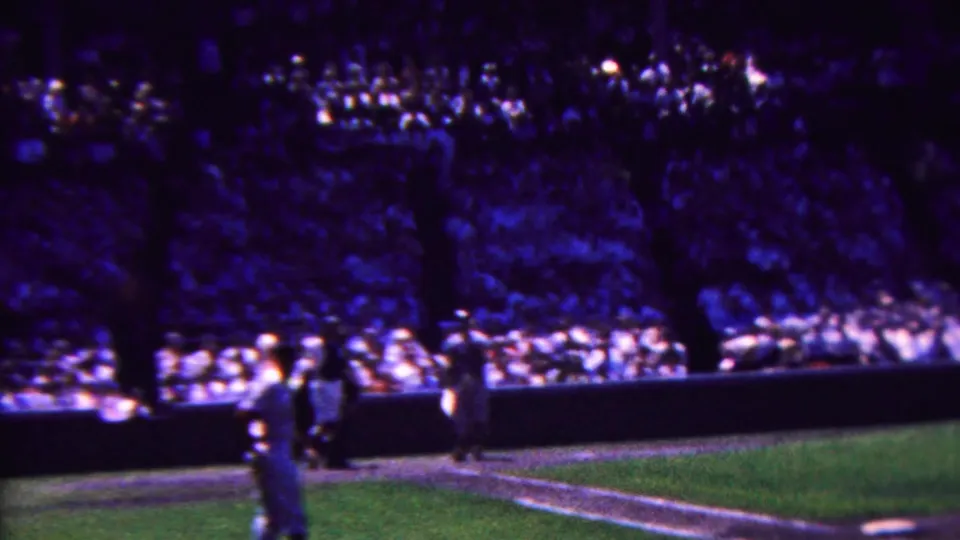 1963 Indiana 8mm home-movie frame showing a crowded baseball stadium, a uniformed player near the field edge, grass, low barriers, and dense spectator seating.