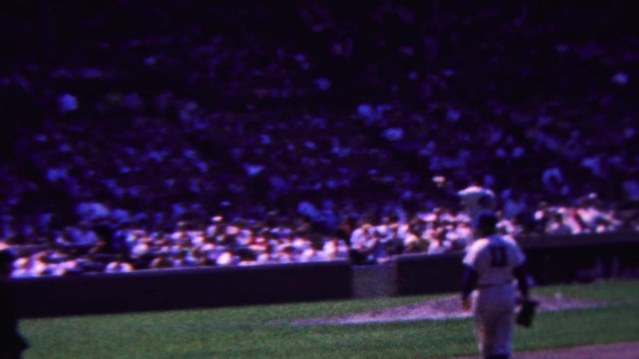 1963 Indiana home-movie view of a baseball field corner with a uniformed player at right, the infield edge, and a crowded stadium beyond.
