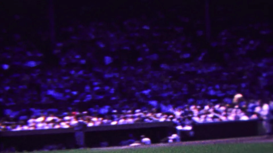 Grainy 1963 Indiana 8mm frame showing a darkened view across a crowded baseball stadium, with the field edge and low barrier at the bottom.