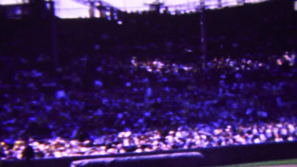 1963 Indiana 8mm home-movie frame looking across a packed baseball stadium, with the field edge, low wall, and bright spectator seating visible through heavy grain.