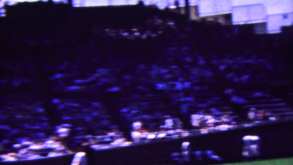 Soft, color-shifted 8mm frame of a crowded baseball stadium in Indiana in 1963, with the field edge and spectator seating visible in daylight.