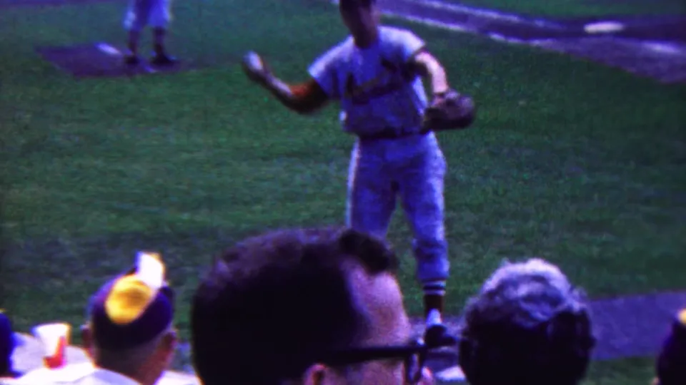 A white-uniformed baseball player appears to gesture toward the crowd from the field in this 1963 Indiana 8mm stadium scene, with spectators visible in the foreground.