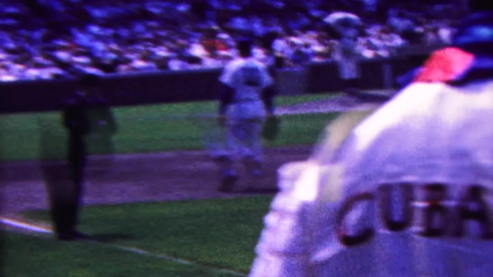 Handheld 8mm frame showing baseball action near home plate, with a white CUBA uniform in the foreground and players and an umpire-like figure beyond.