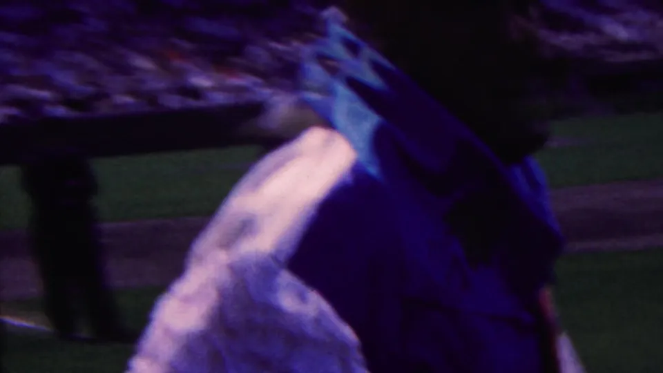 In 1963 Indiana 8mm footage, a close foreground figure partly blocks a baseball field while players and a crowd are visible beyond.
