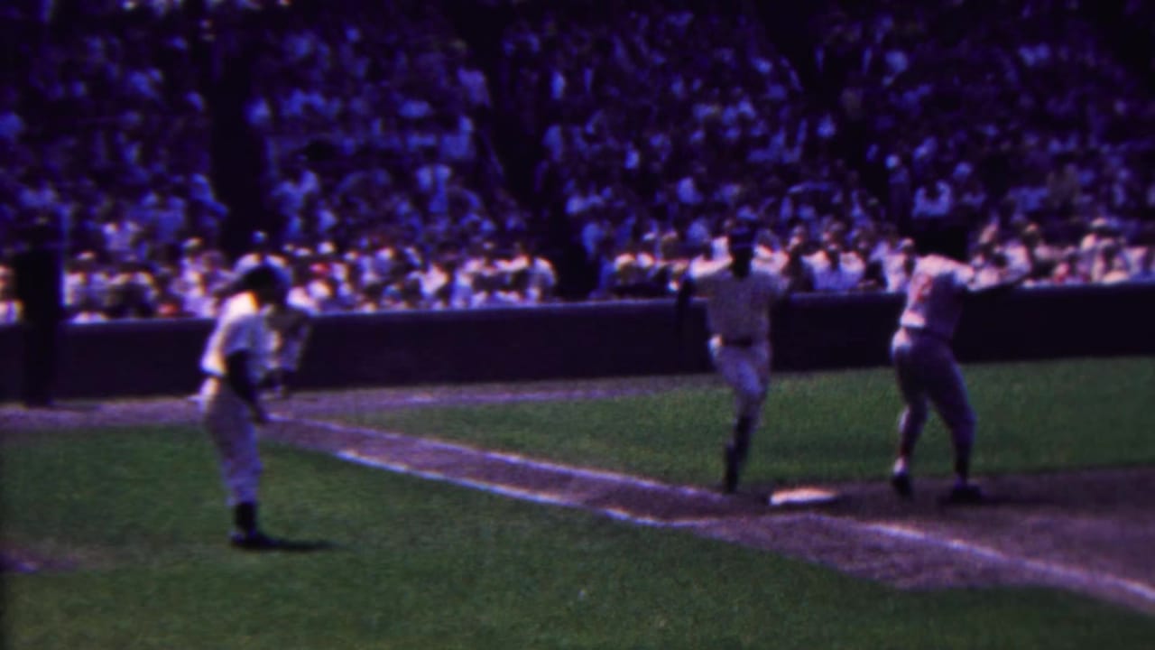 In 1963 Indiana, a baseball runner moves toward or past first base while fielders stand nearby and a packed crowd fills the background.