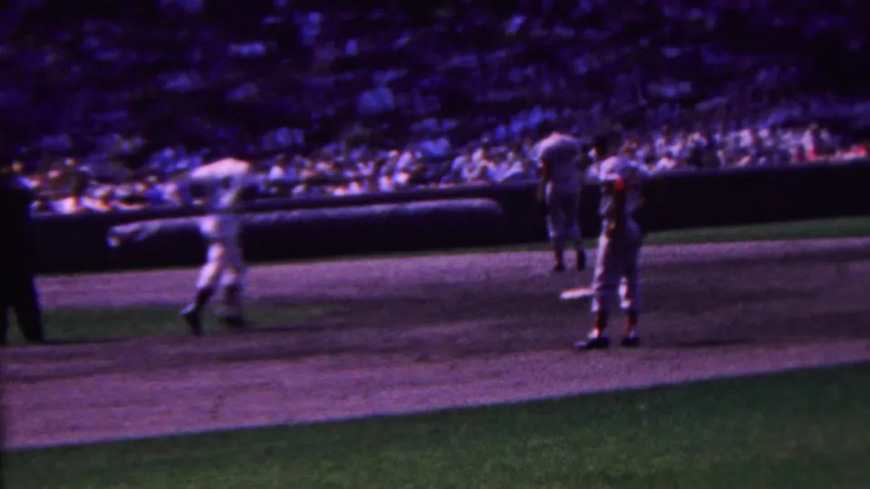 Players in white uniforms are visible near the infield during a 1963 Indiana baseball game, with a distant crowd and blurred field action behind them.