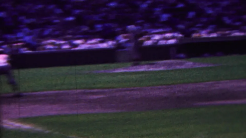 The handheld 8mm frame shows a blurred view across the baseball field in Indiana, with grass, dirt baseline, distant stands, and indistinct players during play.