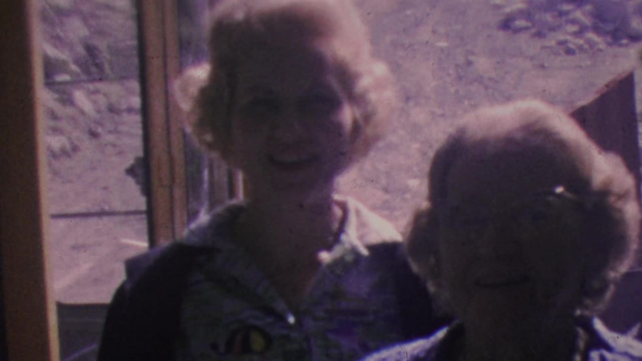 In 1964 Palm Springs Aerial Tramway home-movie footage, a blonde woman in a patterned blouse and dark cardigan stands near another adult beside an exterior doorway, with rocky terrain and tramway structures behind them.