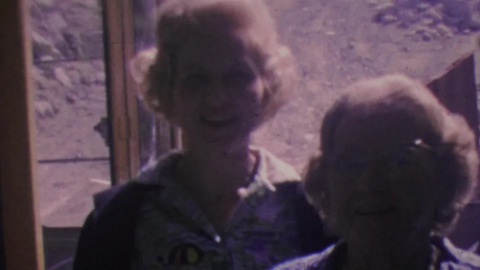 In 1964 Palm Springs Aerial Tramway home-movie footage, a blonde woman in a patterned blouse and dark cardigan stands near another adult beside an exterior doorway, with rocky terrain and tramway structures behind them.