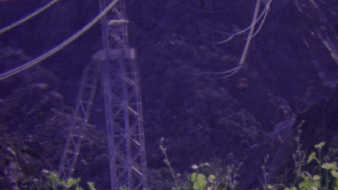 A tramway support tower and overhead cables stand against a dark, rocky mountain slope in the Palm Springs Aerial Tramway setting shown on purple-tinted 8mm film.