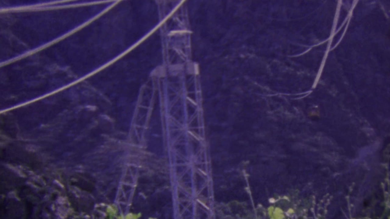 1964 Palm Springs aerial tramway footage showing a steel support tower, suspended cables, and a small tram car against a purple-tinted mountain backdrop.