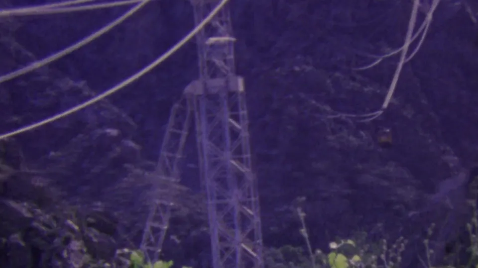 1964 Palm Springs aerial tramway footage showing a steel support tower, suspended cables, and a small tram car against a purple-tinted mountain backdrop.