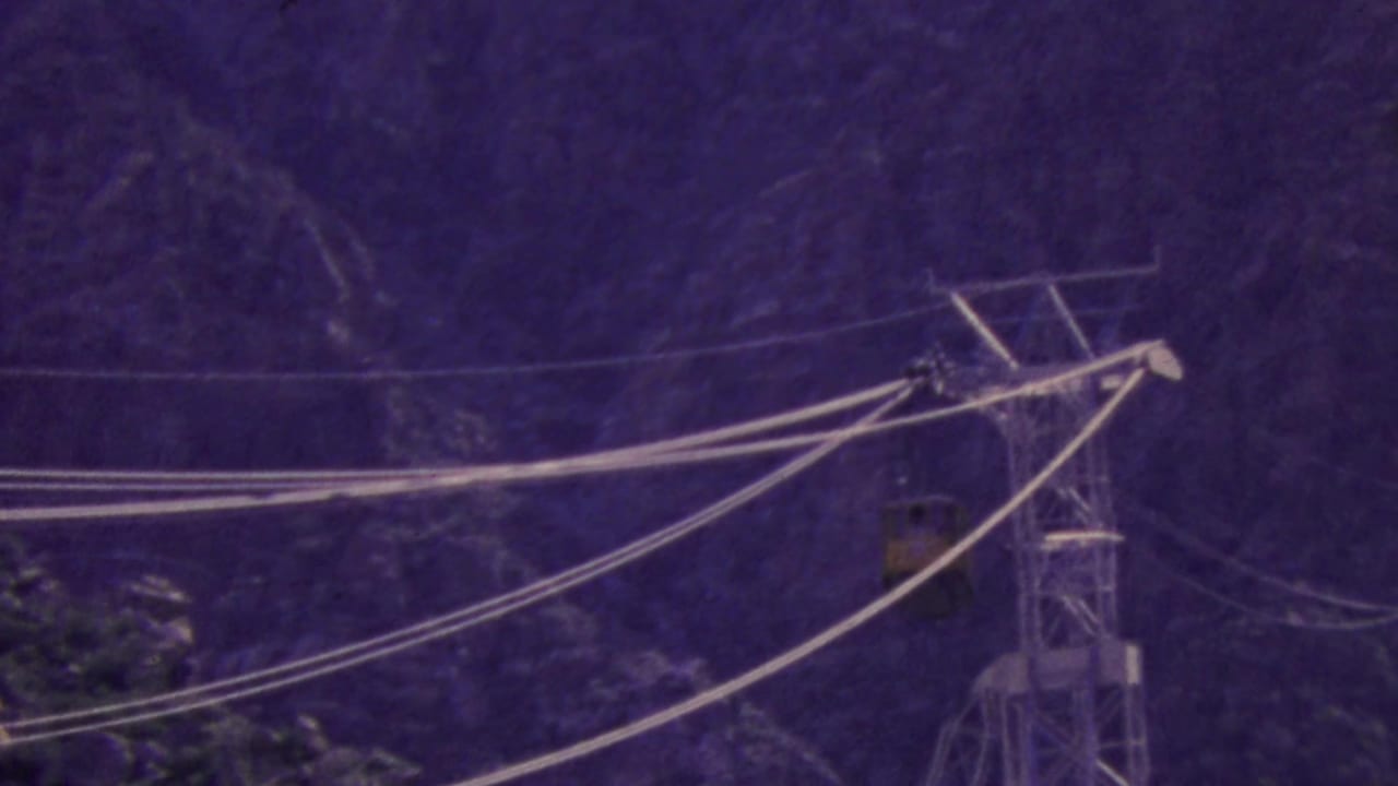 1964 Palm Springs Aerial Tramway view with a yellow tram car crossing steel cables near a support tower on a rugged mountain backdrop.