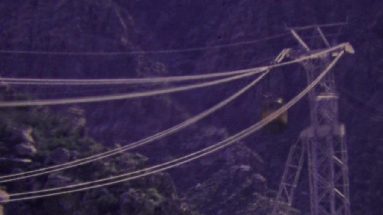 Palm Springs aerial tram cables span the rocky mountain scene, with a support tower visible at right and the small tram car partly obscured by distance and grain.
