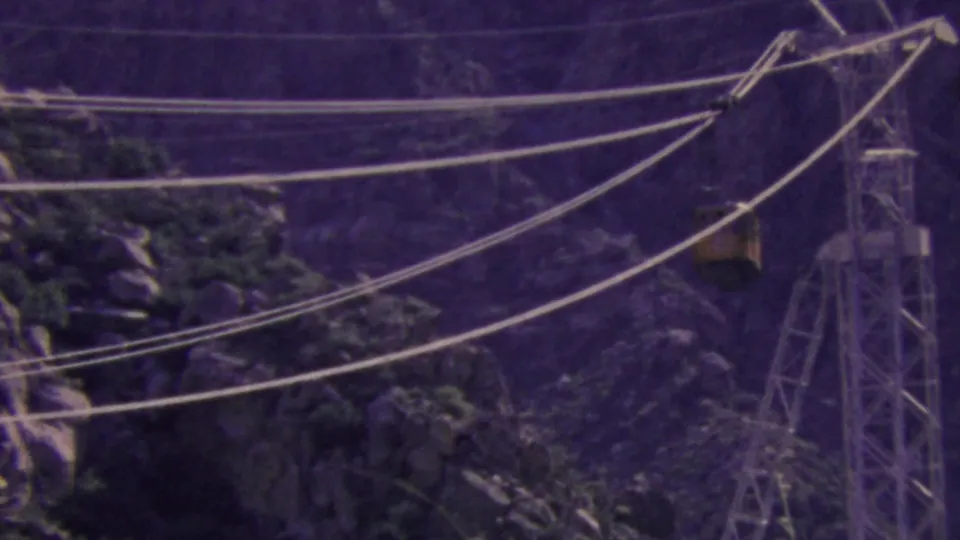 A yellow Palm Springs tram car moves along suspended cables near a lattice support tower, seen through soft 8mm grain and a purple-tinted mountain background.