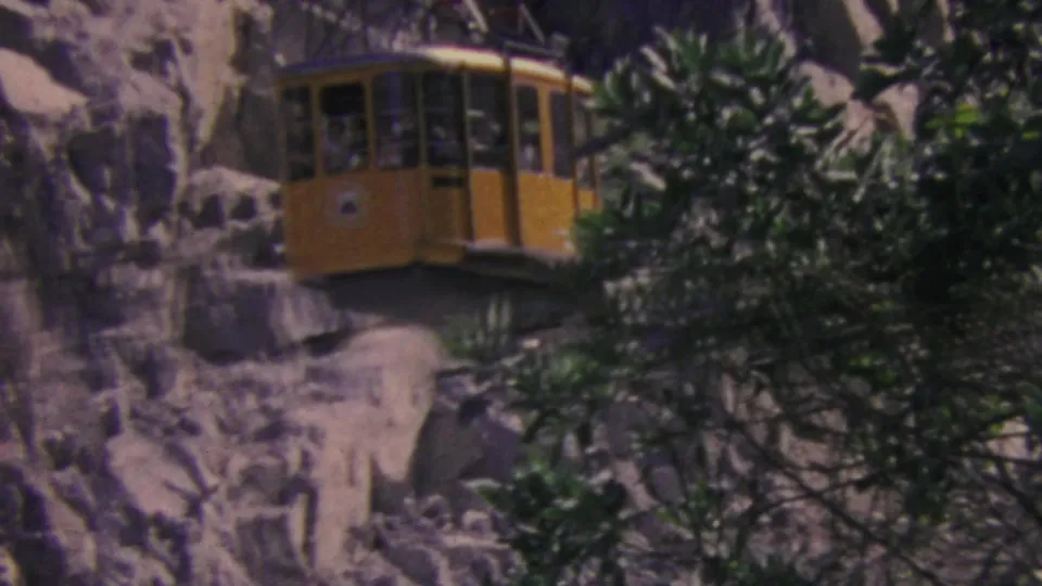Closer 1964 view of a yellow Palm Springs Aerial Tramway car gliding past rock cliffs and evergreen branches, with passengers possibly visible through the windows.