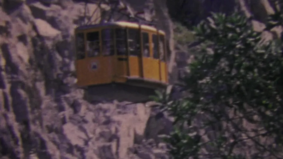 Yellow aerial tram cabin on the Palm Springs mountain route in 1964, seen against pale rocky cliffs with dark green trees partly crossing the frame.