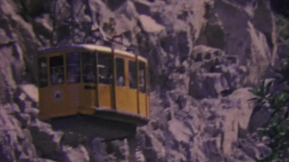 Closer 1964 home-movie view of the yellow aerial tram cabin against pale Palm Springs rock formations, with trees and brush at the edge of the frame.
