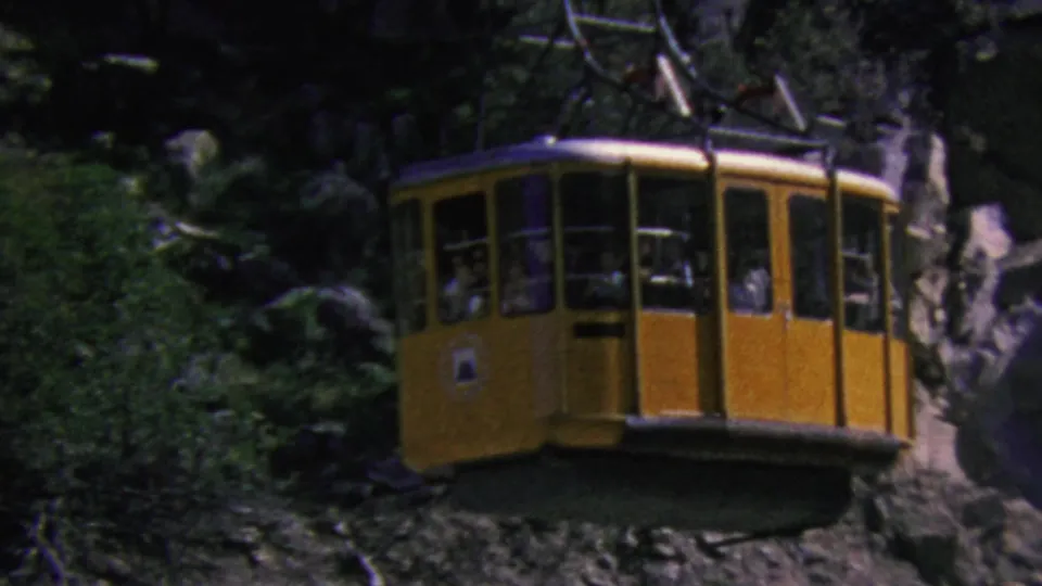 Yellow Palm Springs aerial tram cabin in 1964, partially framed by trees and darker cliff shadows, with passengers faintly visible through the windows.