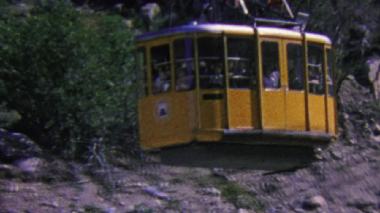 A yellow Palm Springs aerial tramway cabin passes above a rugged mountain slope, with small passenger figures visible through the window openings.