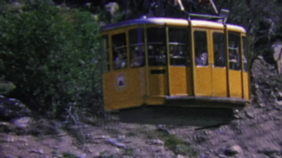 A yellow Palm Springs aerial tramway cabin passes above a rugged mountain slope, with small passenger figures visible through the window openings.