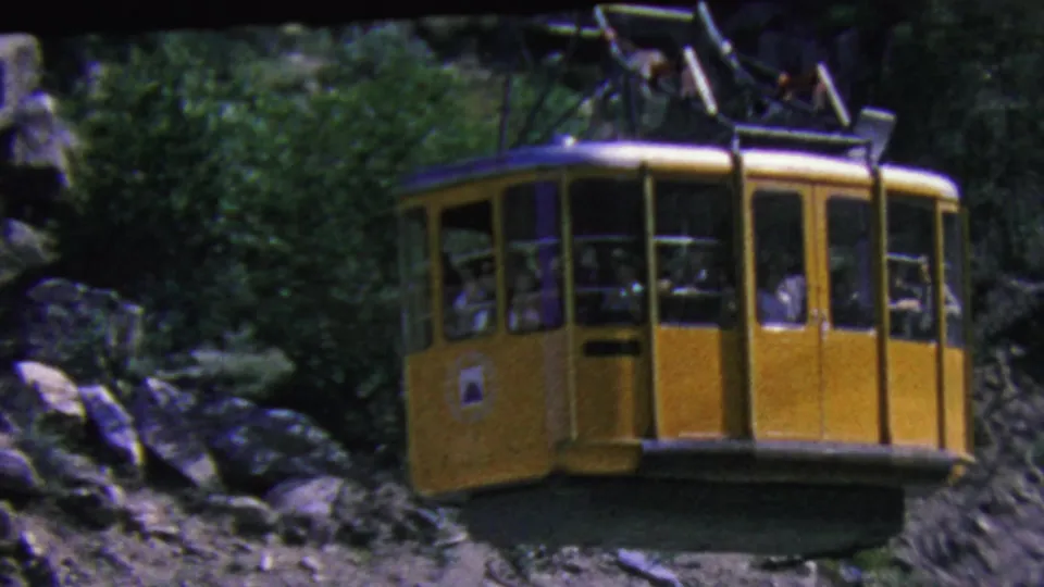 The yellow aerial tram cabin continues across the Palm Springs mountain slope, framed by scrub vegetation, boulders, and the overhead tram cable assembly.