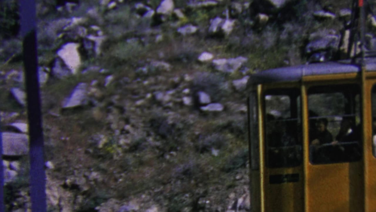 The yellow gondola moves along the Palm Springs mountain cable route, with riders visible as silhouettes inside the cabin and scrub-covered rocks filling the background.