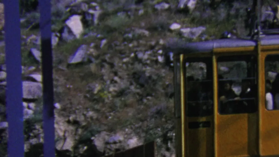 A yellow aerial tramway car is seen lower in the frame, with several passengers visible through the windows as it passes blue support poles on the rocky Palm Springs slope.