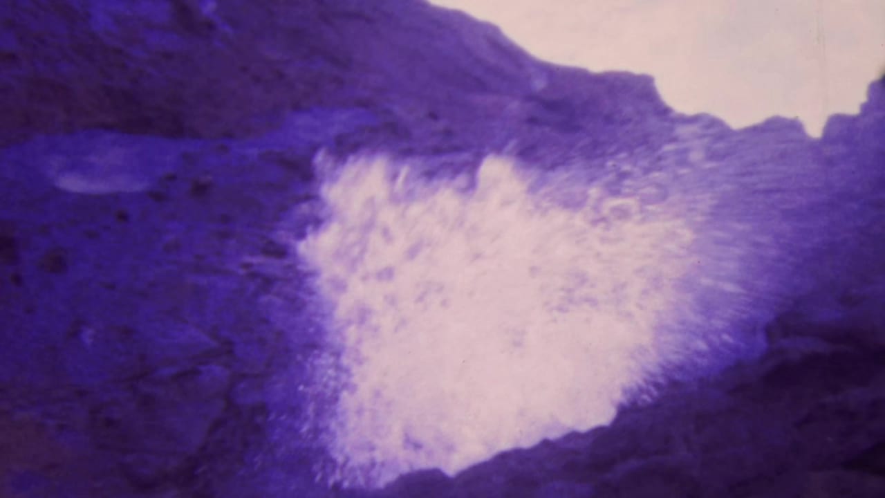 White spray or foam bursts against dark purple rock in a rugged coastal setting.