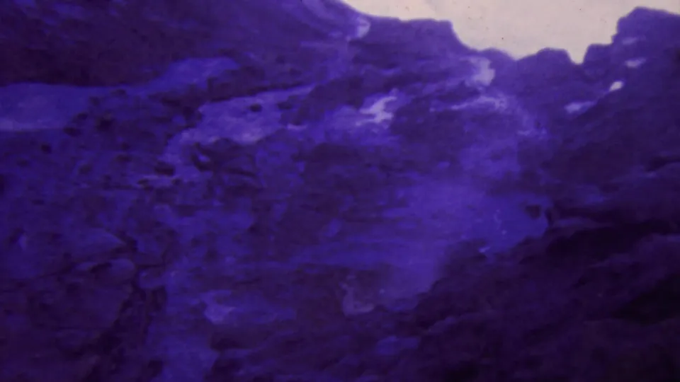 1964 Honolulu-area 8mm frame showing rugged dark rock with a pale wet area or pool, rendered with heavy purple-blue color shift.