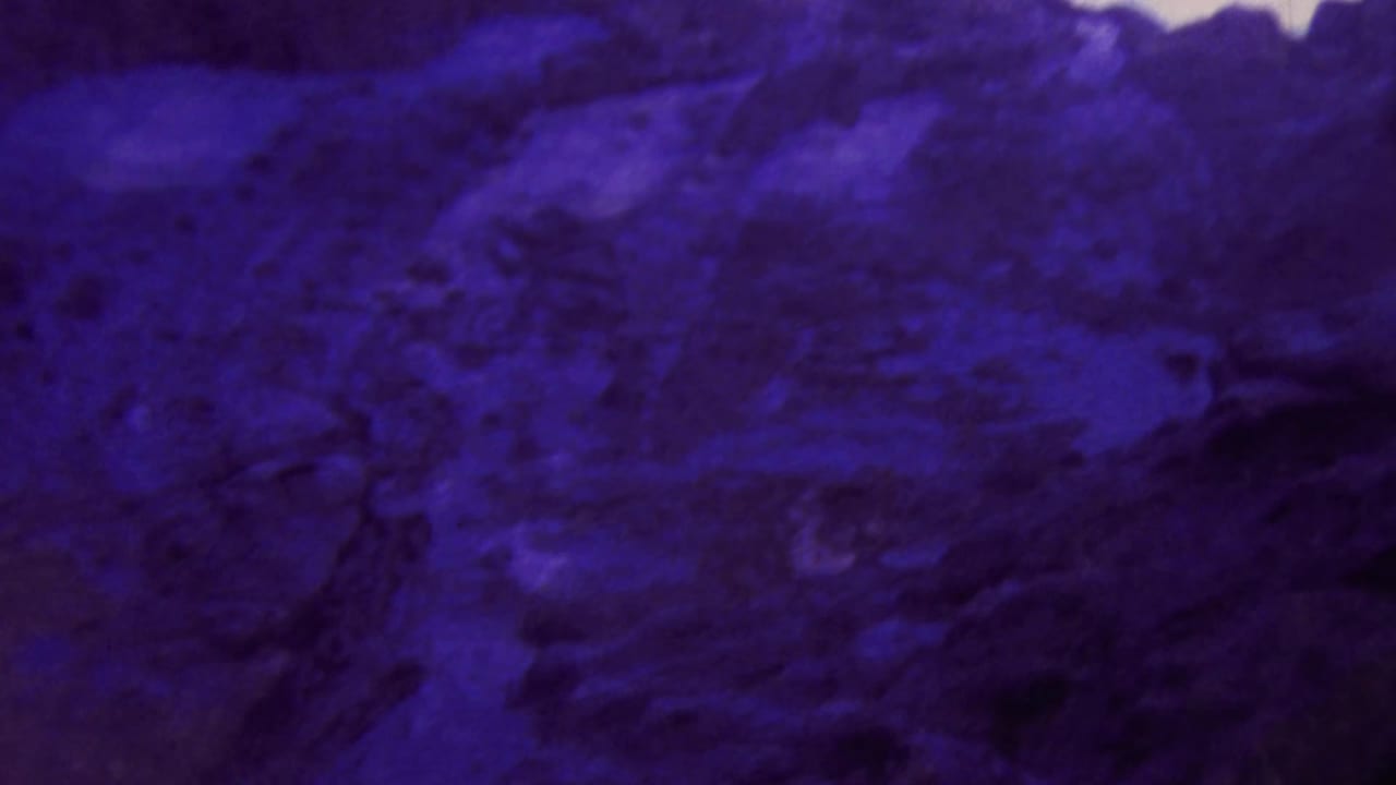 1964 Honolulu home-movie frame showing dark volcanic-looking rock tinted deep blue-purple, with a narrow band of pale sky above.