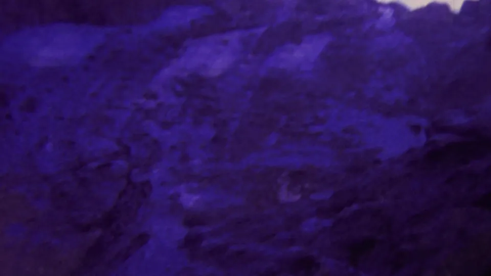 1964 Honolulu home-movie frame showing dark volcanic-looking rock tinted deep blue-purple, with a narrow band of pale sky above.