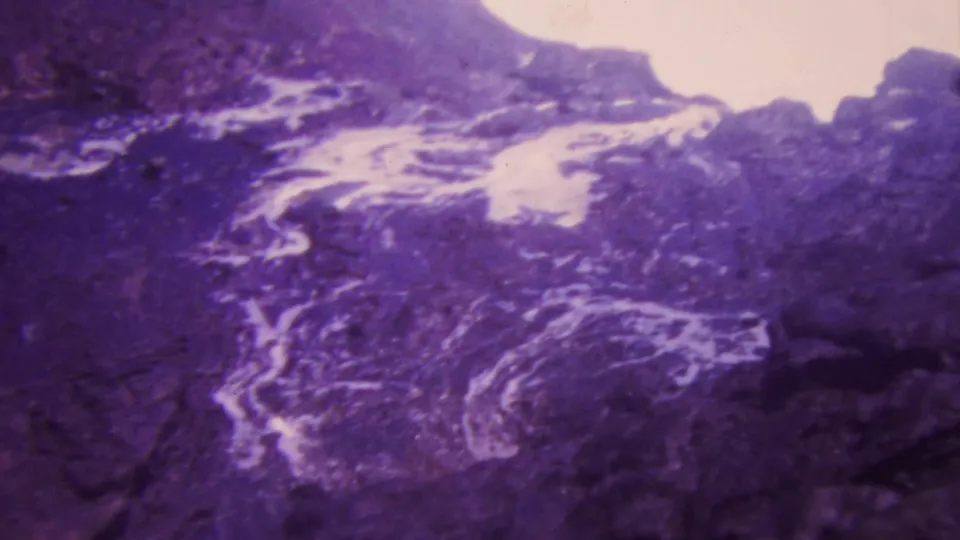 1964 Honolulu home-movie frame showing foaming white water flowing over rugged dark rock with strong purple-blue film coloration.