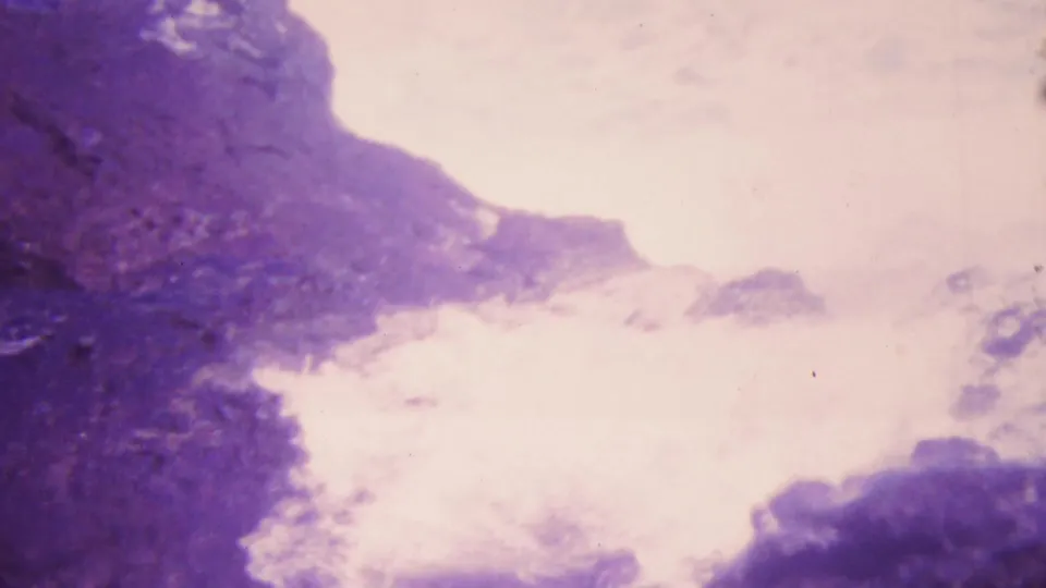 1964 Honolulu 8mm frame with a bright white burst of water, foam, or mist rising beside dark volcanic-looking rock under a pale sky.
